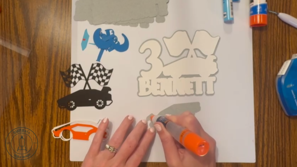 Layering cardstock details onto a cake topper including letters, numbers, and car shapes.